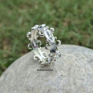 May include: A silver ring with a repeating fleur-de-lis design. The ring is made of a shiny metal and is set against a blurred green background. The ring is on a gray stone. The text "RudraJewelsArt" is visible.