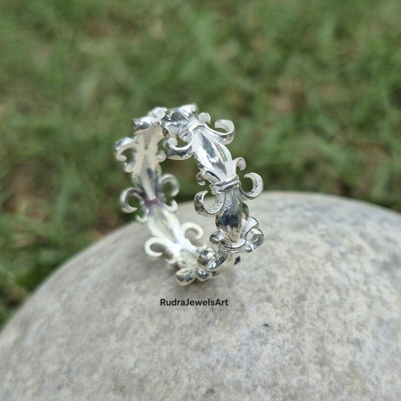 May include: A silver ring with a repeating fleur-de-lis design. The ring is made of a shiny metal and is set against a blurred green background. The ring is on a gray stone. The text "RudraJewelsArt" is visible.