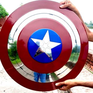 Op de afbeelding: Een Captain America-schild, met een rode buitenring, een zilveren binnenring en een blauw midden met een witte ster. Het schild wordt door twee handen vastgehouden.