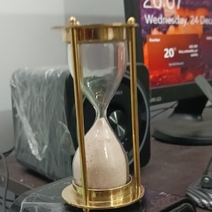 May include: An hourglass with a gold-colored metal frame and base, filled with beige sand. The glass is clear, and the hourglass is set on a black speaker. The background includes a computer monitor displaying the time.