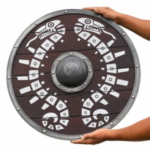 Könnte beinhalten: Ein runder Holzschild mit dunkelbraunem Finish, ausgestattet mit einem zentralen grauen Metallbuckel und einem silbernen Metallrand. Die Oberfläche ist mit zwei symmetrischen, weiß bemalten Schlangenmotiven verziert, die den Buckel umgeben.
