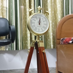 May include: A vintage-style clock with a gold-colored frame and Roman numerals, mounted on a dark brown wooden tripod. The clock face reads "London". The tripod has brass accents and is approximately 40 inches tall.