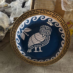May include: A round, decorative shield with a blue center featuring a white owl illustration. The shield has a gold border with black geometric patterns and small gold studs. White wave-like designs and a white olive branch surround the owl.