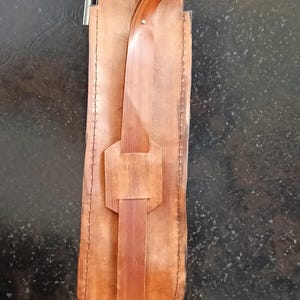 Peut inclure: Un &eacute;tui &agrave; couteau en cuir fait &agrave; la main, de couleur marron clair, avec un design unique. Il est dot&eacute; d'une fermeture &agrave; sangle et boucle, et d'un manche en bois brun plus fonc&eacute;. L'&eacute;tui est con&ccedil;u pour contenir un couteau.