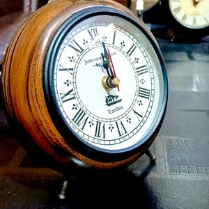 Pode incluir: Um relógio redondo de estilo vintage com caixa de madeira e mostrador branco. O relógio apresenta algarismos romanos pretos, uma moldura preta e o texto "Jefferson Smith London". O relógio tem um pequeno suporte de metal e uma argola no topo.