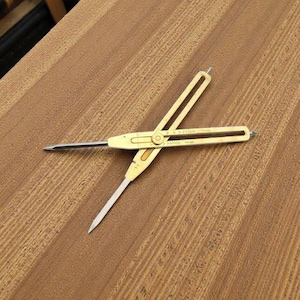 Puede incluir: Un compás de color latón con puntas afiladas, sobre una superficie de madera. La herramienta tiene una bisagra en el centro y se utiliza para medir o transferir distancias. La veta de la madera es visible en el fondo.