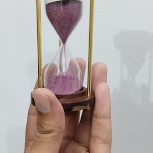 Könnte beinhalten: Eine Sanduhr mit Holzrahmen und messingfarbenen Metallstützen. Der Sand im Inneren hat eine tiefviolette Farbe. Die Sanduhr wird in einer Hand gehalten.