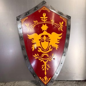 May include: A pointed heater shield featuring a deep red surface with a yellow graphic emblem of a central crest flanked by stylized wings and floral motifs. The shield has a metallic silver border with visible rivets along the outer edge.