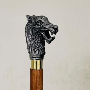 Könnte beinhalten: Ein Spazierstock mit einem Wolfskopfgriff. Der Griff ist ein detaillierter, silberfarbener Wolfskopf mit offenem Maul. Der Stock hat einen Messingkragen und einen Holzschaft. Das ist ein dekoratives Accessoire.