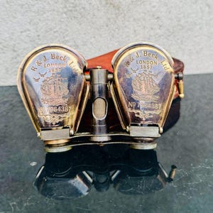 May include: Antique brass binoculars with a leather case. The binoculars are engraved with "R & J. Beck, London 1857" and a ship illustration. The serial number "No 7964387" is also visible. The binoculars have a vintage aesthetic.