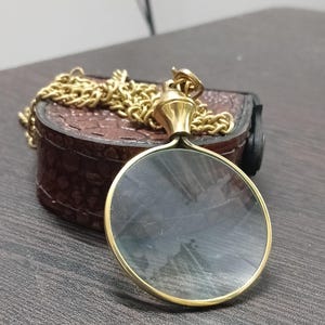 Puede incluir: Una lupa dorada con una cadena, apoyada sobre un estuche de cuero marrón. La lupa tiene una lente circular y un asa decorativa. La cadena es de estilo de eslabones clásico.