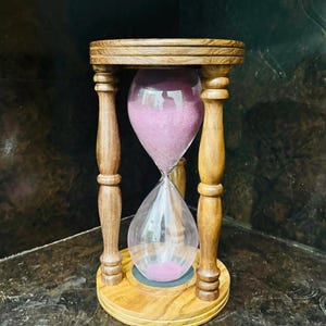May include: An hourglass with a wooden frame and base. The hourglass has a clear glass bulb filled with pink sand. The wooden frame is a light brown colour. The hourglass is a decorative item.