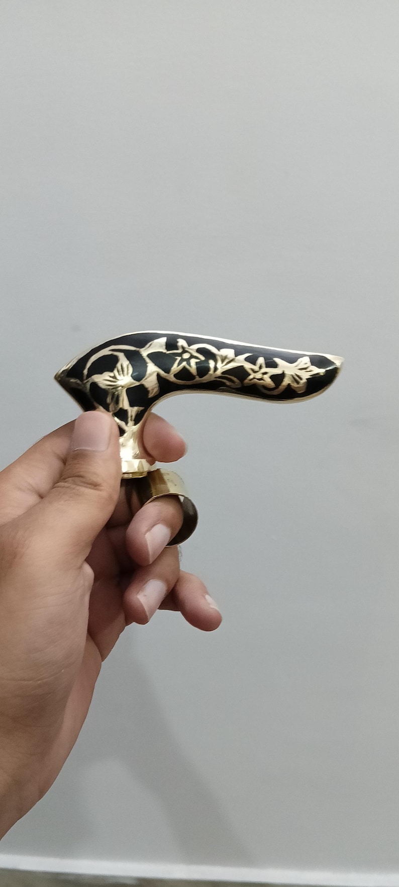 Pu&ograve; includere: Maniglia in ottone ornata con un motivo floreale nero. La maniglia presenta una forma curva e un anello per un dito. Il design &egrave; probabilmente per un bastone o un bastone da passeggio.