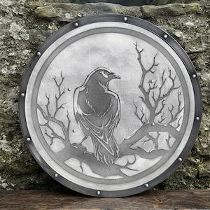 May include: A circular metal shield featuring a detailed engraved design of a raven perched on a bare tree branch. The shield has a dark, riveted outer rim and a brushed silver-toned centre, showcasing a monochromatic aesthetic with clean lines.
