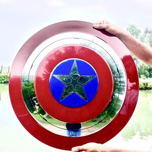 Op de afbeelding: Een rood, wit en blauw schild met een ster in het midden. Het schild heeft een cirkelvormig ontwerp met een zilveren rand. Het schild wordt omhoog gehouden tegen een achtergrond van water en lucht. De diameter is ongeveer 76 cm.