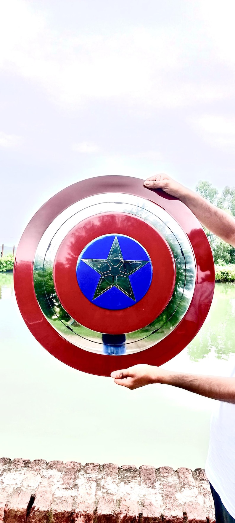 Op de afbeelding: Een rood, wit en blauw schild met een ster in het midden. Het schild heeft een cirkelvormig ontwerp met een zilveren rand. Het schild wordt omhoog gehouden tegen een achtergrond van water en lucht. De diameter is ongeveer 76 cm.