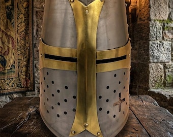 May include: A silver-colored medieval Crusader helmet with a gold-colored cross design on the front. The helmet has small ventilation holes and is set against a rustic background, suggesting historical or fantasy themes.