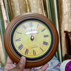 May include: A round wooden clock with a yellow face and black Roman numerals. The clock has a black bezel and the text "1747 LONDON" is visible. The clock is being held by a person.