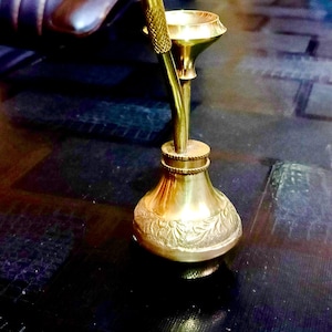 May include: A small, ornate brass hookah with a bulbous base and a slender, curved stem. The base features a detailed floral pattern. The hookah is designed for smoking and sits on a dark, reflective surface.