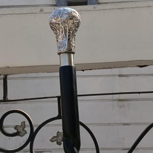 Może przedstawiać: Dekoracyjna laska do chodzenia ze srebrnym, ozdobnym uchwytem. Uchwyt ma szczegółowy kwiatowy wzór. Trzon jest czarny i ma skręcony wzór liny. Laska to stylowy dodatek.