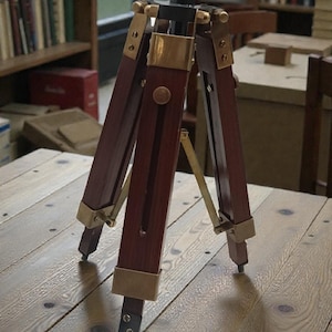 May include: A vintage wooden tripod with brass accents. The tripod has three legs and a dark wood finish. The brass details include the leg joints and the top mounting plate. The tripod is set on a wooden table.