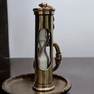 May include: A brass hourglass with a decorative design, set on a matching brass base. The hourglass is encased in a cylindrical frame with an oval window, and the sand inside is white. The piece has a vintage aesthetic.