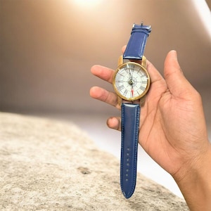 May include: A vintage-style wristwatch with a gold-tone case and a white face featuring black Roman numerals and red accents. The watch has a blue leather band. The watch is held against a neutral background.