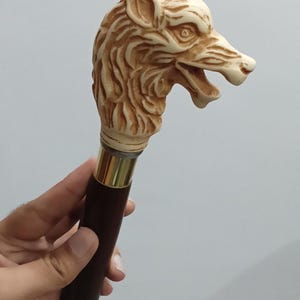 Puede incluir: Bastón con un mango de cabeza de lobo tallada. La cabeza de lobo es blanquecina con detalles de pelaje y rasgos faciales. El eje es de color marrón oscuro con una banda metálica dorada cerca del mango.