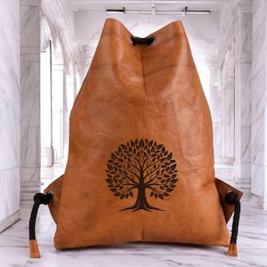 May include: A tan drawstring backpack featuring a dark brown tree graphic centered on the front. The bag is constructed from a smooth, leather-like material with black braided rope cords threaded through metal eyelets at the top corners.