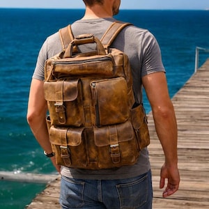 May include: A brown leather backpack with several pockets and compartments. The bag features a top handle, adjustable shoulder straps, and buckle fastenings. The backpack is worn by a person on a wooden pier, with the sea visible in the background.