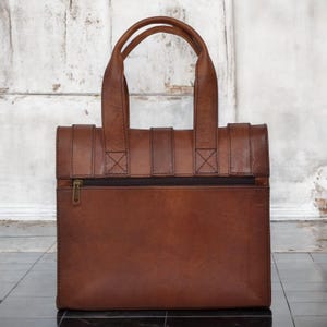 May include: A brown leather tote bag with two top handles and a zippered front pocket. The bag has a rectangular shape with a structured design and visible stitching details. The bag is made of a rich, textured leather.