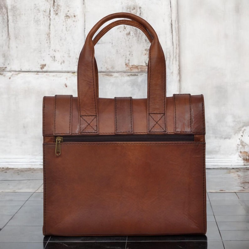 May include: A brown leather tote bag with two top handles and a zippered front pocket. The bag has a rectangular shape with a structured design and visible stitching details. The bag is made of a rich, textured leather.