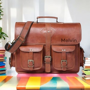 May include: A brown leather satchel with two front pockets, a top handle, and a shoulder strap. The bag has a buckle closure and the name "Melvin" is printed on the front. The bag is sitting on a colorful table.