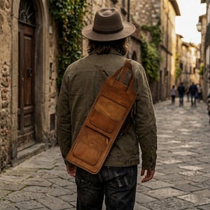 Op de afbeelding: Een persoon met een bruine fedora-hoed en een olijfgroene jas loopt over een geplaveide straat. Een bruine leren schoudertas met meerdere vakken hangt over de schouder. De tas lijkt handgemaakt.