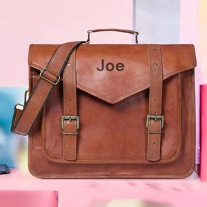 May include: A brown leather satchel with a top handle and adjustable shoulder strap. The bag features two front straps with buckles and the name "Joe" printed on the front. The bag is sitting on a pink surface.
