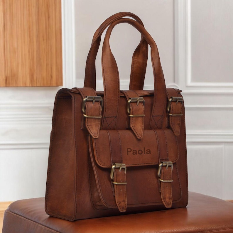 May include: A brown leather satchel bag with two top handles and a front flap pocket. The bag features multiple buckles and the name "Paola" embossed on the front. The bag is sitting on a brown surface.