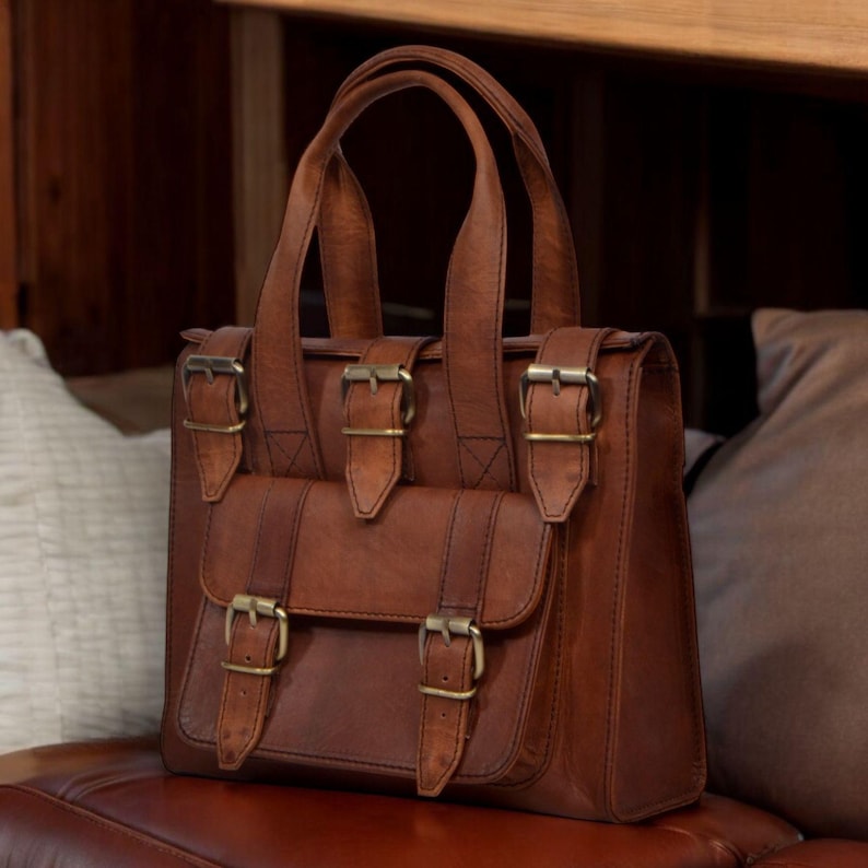 May include: A brown leather handbag with a top handle and a front flap pocket. The bag features brass-colored buckles and hardware. The bag is sitting on a brown leather surface.