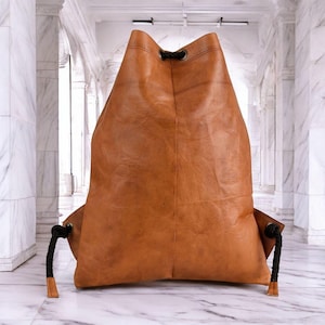 Peut inclure: Un sac à dos marron en cuir avec des cordons noirs. Le sac a une forme arrondie et une surface texturée. L'arrière-plan est un couloir en marbre.
