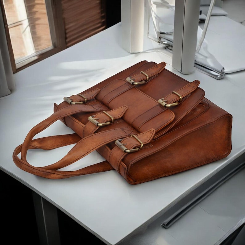 May include: A brown leather satchel with multiple brass buckles and a top handle. The bag is rectangular with a flap closure and two straps across the front. The bag is sitting on a white surface.