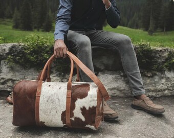 May include: A brown and white cowhide duffel bag with brown leather handles and straps. The bag has a rectangular shape and is being held by a person. The background features a natural outdoor setting with trees and greenery.