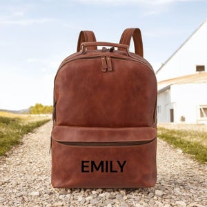 Könnte beinhalten: Brauner Lederrucksack mit dem Namen "EMILY" in Schwarz auf der Vordertasche aufgedruckt. Der Rucksack hat zwei Schultergurte, einen oberen Griff und mehrere Reißverschlussfächer. Die Tasche steht auf einem Schotterweg.