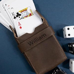 May include: A brown leather card case with the name "William" embossed on the strap. The case holds a deck of playing cards, with the King of Clubs and Ace of Diamonds visible. Dice are scattered around the case on a blue surface.