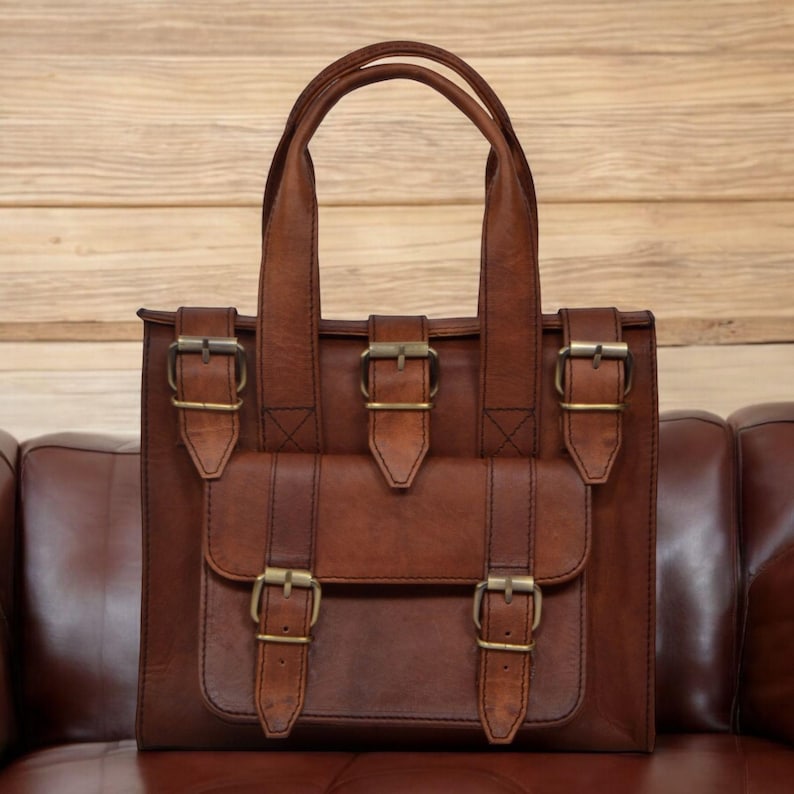 May include: A brown leather tote bag with two top handles and a front flap pocket. The bag features multiple brass-colored buckles and a structured design. The bag is set against a wooden background.