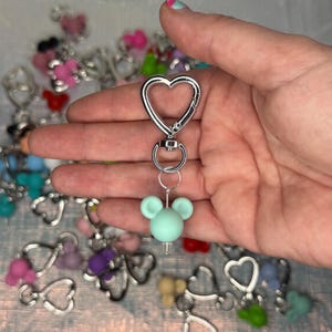 May include: A mint green silicone bead shaped like a mouse head with two round ears, attached to a silver-colored metal heart-shaped lobster clasp keychain. The keychain features a circular ring connector between the clasp and the bead charm.