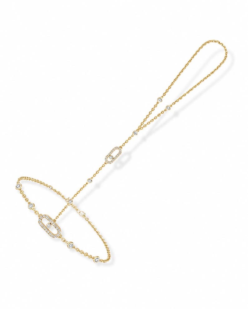 May include: A gold hand chain bracelet featuring a delicate chain design with two oval-shaped links encrusted with small clear stones. The piece includes additional round bezel-set stones spaced along the chain, connecting a ring loop to a wrist band.