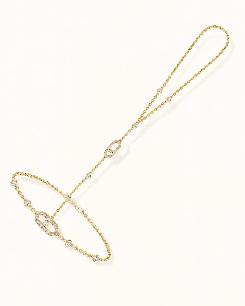 May include: A gold hand chain bracelet featuring a delicate chain adorned with small round clear stones. The design includes two oval-shaped links encrusted with clear stones, one positioned on the wrist section and one on the finger loop.