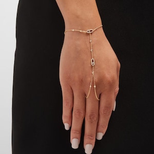 May include: A gold hand chain bracelet featuring delicate link chains adorned with small round diamonds and two elongated oval-shaped charms, each set with a central diamond, connecting the wrist section to a ring loop worn on the middle finger.