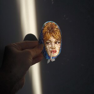 May include: A beaded brooch featuring a portrait of a woman with blonde hair and a blue head covering. The face is detailed with red lips and light-colored skin. The brooch is oval-shaped and held against a dark background.