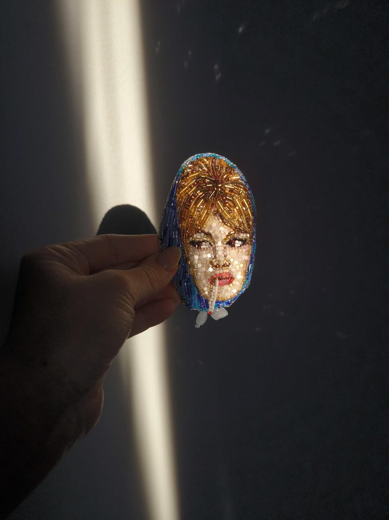 May include: A beaded brooch featuring a portrait of a woman with blonde hair and a blue head covering. The face is detailed with red lips and light-colored skin. The brooch is oval-shaped and held against a dark background.