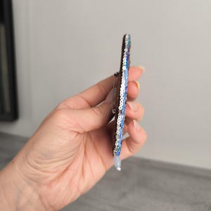 May include: A handmade brooch with a dark brown base, adorned with blue and silver sequins. The brooch has a pin on the back for easy attachment. The design features a gradient of blue sequins, creating a shimmering effect.